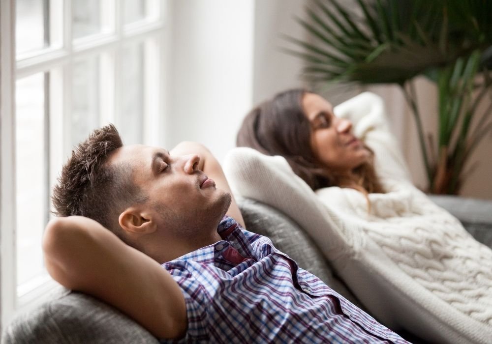 A couple relaxing comfortably on a sofa with their eyes closed, enjoying the cool indoor climate of their home.
