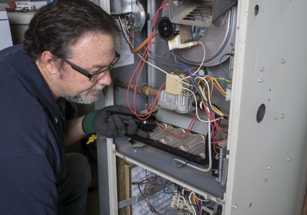 An HVAC technician using a flashlight to perform a detailed inspection of the burners and heat exchanger inside a residential furnace.